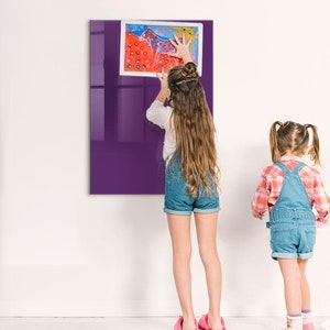 Op de afbeelding: Twee kinderen, waarvan er één op een ladder staat, hangen een kleurrijk schilderij op een paarse magnetische whiteboard. Het bord is gemonteerd op een witte muur.
