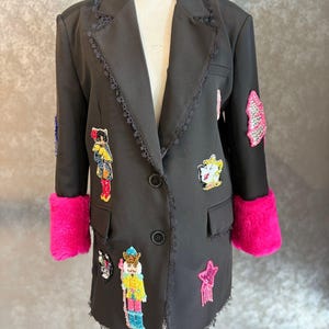 Può includere: Blazer nero con polsini in finta pelliccia rosa acceso e toppe decorative. Il blazer ha un bordo in pizzo. Le toppe includono una stella, un gatto e un soldato.