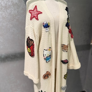 Könnte beinhalten: Cremefarbene Strickjacke mit offenem Frontdesign, verziert mit bunten, gestickten Aufnähern. Zu den Aufnähern gehören ein rot-weiß karierter Stern, eine Zeichentrickfigur, eine Pommes-Frites-Box und ein Hello Kitty-Design. Der Pullover hat lange Ärmel und eine lockere Passform.