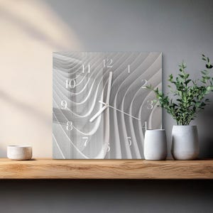Puede incluir: Reloj de pared cuadrado con un diseño moderno. La esfera del reloj presenta un patrón abstracto gris y blanco con números y manecillas blancos. El reloj se exhibe en una estantería de madera con objetos decorativos.