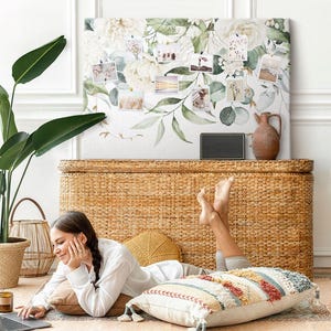 May include: A rectangular canvas wall art piece featuring a printed design of white flowers and green eucalyptus leaves. The canvas serves as a display board with several small photographs attached to its surface, showcasing a floral botanical theme.