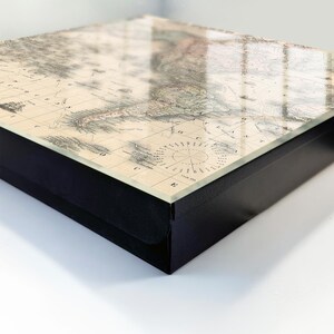 Può includere: Una scatola quadrata con cornice nera e superficie superiore lucida che presenta una mappa in stile vintage del Sud America. La mappa include linee di griglia dettagliate, etichette geografiche e un design a rosa dei venti circolare.