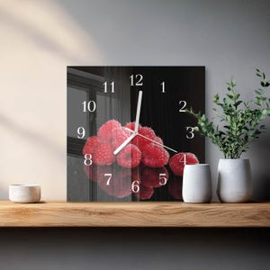 May include: Square wall clock with a black background featuring a cluster of red raspberries. White clock hands and numerals are visible. The clock is displayed on a wooden shelf with decorative items.