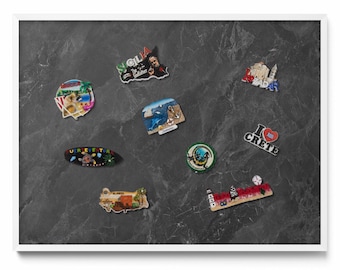 May include: A collection of souvenir magnets with various designs, including landmarks, maps, and text. The magnets are in different shapes and colors, with text such as "Sicilia" and "I Love Crete". The magnets are displayed on a dark gray surface.
