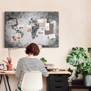 May include: A woman sits at a desk with a world map corkboard above her. The corkboard has photos and notes pinned to it. The desk is made of wood and has a black metal frame. There is a potted plant on the desk and a potted plant on a black side table.