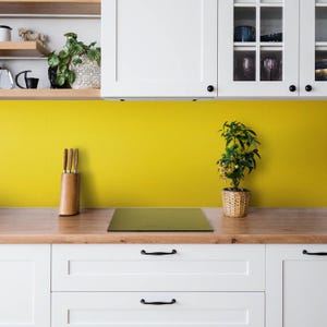 May include: A kitchen backsplash featuring a solid mustard yellow color, installed behind a wooden countertop with a wooden knife block, a small potted green plant, and white cabinetry above and below the workspace.