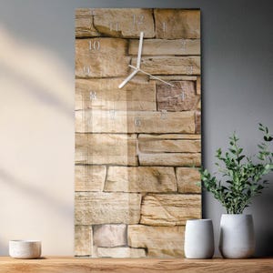 Op de afbeelding: Een rechthoekige wandklok met een stenen baksteenontwerp. De wijzerplaat van de klok heeft zilveren cijfers en wijzers. De achtergrond is een textuurpatroon van beige en bruine stenen, wat een rustieke esthetiek creëert.