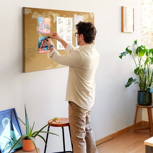 May include: A person adds a colorful illustration to a corkboard with a world map design. The corkboard is hanging on a white wall in a home office.
