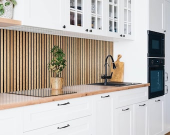 May include: A kitchen featuring white cabinets with black hardware, a light wood countertop, and a vertical wood slat backsplash. A black faucet sits over a black sink, with a small potted plant and a wooden cutting board on the counter.