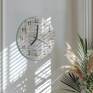 May include: A round wall clock with a light gray stone-like face. Black numerals mark the hours, and black hands indicate the time. The clock is mounted on a white wall, with a glass border.