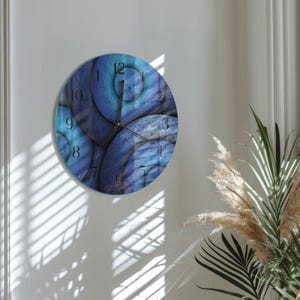 May include: A circular wall clock with an abstract blue design. The clock face has overlapping circular shapes in various shades of blue, with black hands and numerals. The clock is mounted on a white wall, with shadows from a window visible.