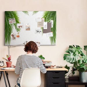 May include: A rectangular white pin board featuring green palm leaf illustrations on the left and right edges. The board displays various pinned photographs, notes, and a card with the text Thank you, mounted on a light-colored wall above a desk.