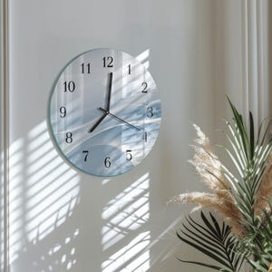 May include: A round wall clock with a clear glass face and black hands. The clock face features a blue and white abstract design. The numbers are black and the clock is mounted on a white wall.