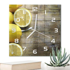 Può includere: Un orologio da parete quadrato con una stampa fotografica di limoni giallo brillante e un limone tagliato su una superficie in legno marrone rustico. Il quadrante mostra numeri arabi bianchi da 1 a 12 e sottili lancette bianche.