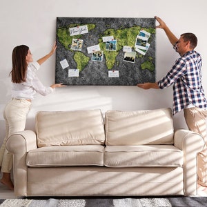 May include: A large, rectangular world map wall decor with green continents and a dark gray background. The map is adorned with photos and pins, and is being hung on a wall by two people.