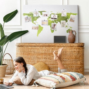 Peut inclure: Une femme se détend sur le sol dans un salon, en utilisant un ordinateur portable. Elle est entourée d'oreillers et d'un coffre en osier. Une carte du monde avec des photos épinglées dessus est accrochée au mur derrière elle.