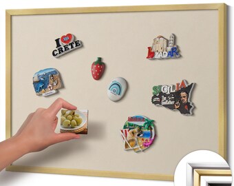 May include: A collection of souvenir magnets on a beige magnetic board with a gold frame. The magnets depict various images, including a strawberry, a dog on a beach, and the words "I Crete". A hand is holding a magnet with olives.