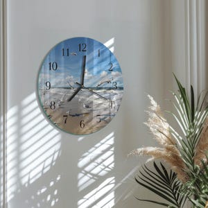 May include: A round wall clock featuring a beach scene with seagulls, blue sky, and white clouds. The clock face displays black numerals and hands. The clock is mounted on a white wall, with shadows from a window visible.
