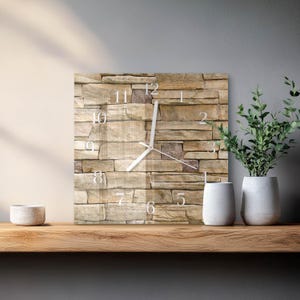 May include: Square wall clock with a stone-like texture background. The clock face features white numerals and hands. The clock is displayed on a wooden shelf, with white vases and greenery to the right and a small bowl to the left.