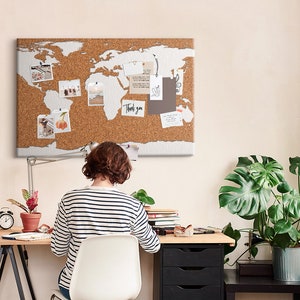 May include: A person sits at a desk with a corkboard world map above them. The corkboard has photos, notes, and other items pinned to it. The desk is made of wood and has a black metal frame. There is a potted plant on the desk and another potted plant on a black side table.