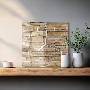 May include: A square wall clock featuring a printed image of stacked tan and brown stones on its face. The clock has white hour, minute, and second hands, and white Arabic numerals from one to twelve positioned over the stone pattern.