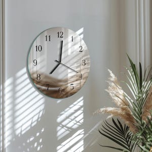 May include: A round wall clock with a clear glass face and black hands and numerals. The clock's face has a subtle, abstract design in shades of brown and beige. The clock is mounted on a white wall.