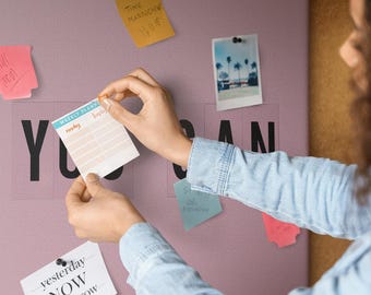 Puede incluir: Un bloc de notas semanal cuadrado con un encabezado azul claro y una cuadrícula para la planificación diaria. El bloc se sostiene contra un tablero de pared malva con letras negras, notas adhesivas de colores y una fotografía.