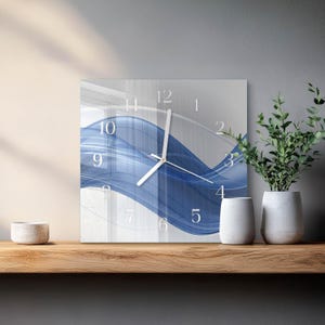 May include: Square wall clock with a blue and white abstract wave design. The clock face features white numerals and hands. The clock is displayed on a wooden shelf with decorative items.