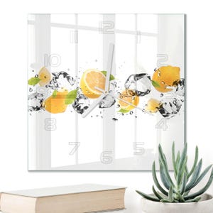 May include: A square wall clock with a white background featuring a graphic of yellow lemon slices, green leaves, and clear ice cubes. The clock face displays large, outlined numbers from one to twelve and has thin white hour and minute hands.
