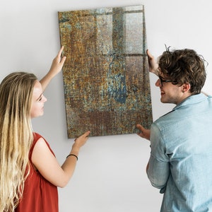 Könnte beinhalten: Zwei Personen hängen ein großes, rechteckiges abstraktes Gemälde an eine weiße Wand. Das Gemälde hat einen rostigen, metallischen Look mit Brauntönen, Blau- und Grüntönen.