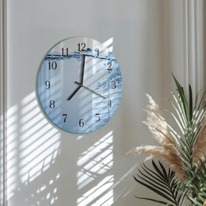 Può includere: Un orologio da parete rotondo con un design a tema acqua. Il quadrante dell'orologio presenta un motivo ad acqua blu e bianco, lancette e numeri neri. L'orologio è montato su una parete bianca, con ombre di persiane visibili.