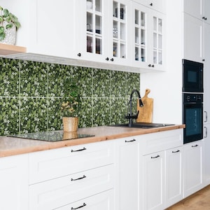 May include: A kitchen backsplash featuring square tiles with a dense botanical pattern of green leaves and small white flowers. The tiles are installed above a light wood countertop, white cabinets, a black faucet, and a black built-in oven.
