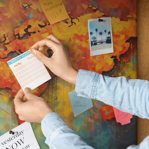 May include: A person is adding a weekly planner to a corkboard with a world map design. The planner has a blue and teal border and the words "Weekly Planner" printed on it. The planner has a column for each day of the week, starting with Monday and ending with Sunday. There are also several other notes and a photo on the corkboard.