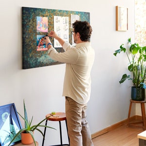 Pode incluir: Uma pessoa adiciona uma ilustração colorida a um quadro de cortiça com um fundo texturizado azul e verde. O quadro de cortiça está pendurado em uma parede branca.