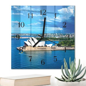 Könnte beinhalten: Quadratische Wanduhr mit einer malerischen Ansicht des Sydney Opera House. Das Zifferblatt zeigt schwarze Ziffern und Zeiger vor dem Hintergrund von blauem Wasser, Himmel und dem ikonischen Gebäude. Die Uhr ist quadratisch und scheint aus Glas zu sein.