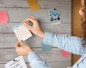 Puede incluir: Un bloc de notas de planificación semanal con encabezado azul claro y líneas de cuadrícula color melocotón, con el texto Weekly Planner y Monday. El bloc se está fijando en una pared de paneles de madera blanca junto a notas adhesivas.