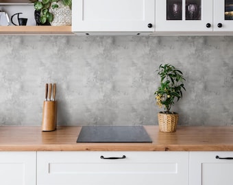 May include: A kitchen scene featuring white cabinets, a wooden worktop, and a grey textured splashback. A wooden knife block with three knives sits on the left, and a small green plant in a woven basket sits on the right of a black hob.