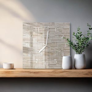 May include: Square wall clock with a light beige stone-like face. The clock has white hands and white numbers. The clock is displayed on a wooden shelf with two white vases and a small white bowl. Green plants are in the vases.