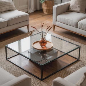 Peut inclure: Une table basse carrée moderne avec un plateau en verre et un cadre en métal foncé. La table a une étagère inférieure en bois. Un petit vase avec des plantes séchées est posé sur un plateau en bois sur la table. Deux canapés blancs sont en arrière-plan.