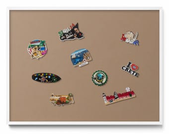 May include: A collection of colorful souvenir magnets, including shapes of islands, landmarks, and phrases like "I Love Crete." The magnets are displayed on a neutral brown surface within a white frame.