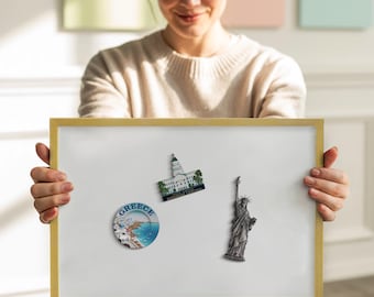 May include: A white framed magnetic board with three travel-themed magnets. The magnets depict the Statue of Liberty, a building with a green roof, and a round magnet with the word "Greece" and a coastal scene.