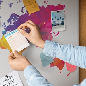 May include: A person is adding a weekly planner to a corkboard with a colorful world map background. The planner has the words "Weekly Planner" and "Monday" printed on it. There are other notes and a photo on the corkboard.