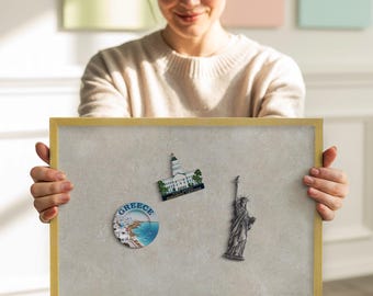 May include: A light beige framed bulletin board displaying travel-themed magnets. The magnets feature the Statue of Liberty, a Greek coastal scene, and a building with a green roof. The frame is a light gold color.