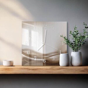 Op de afbeelding: Vierkante wandklok met een neutraal kleurenpalet. De wijzerplaat heeft een zacht, abstract ontwerp met witte cijfers en wijzers. De klok wordt getoond op een houten plank met decoratieve witte vazen en een plant in pot.