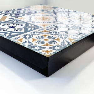 Può includere: Un vassoio decorativo quadrato con cornice nera e superficie superiore lucida caratterizzata da un motivo ripetitivo di disegni geometrici e floreali in blu, bianco e oro. Il vassoio presenta una finitura liscia e riflettente.