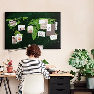 May include: A home office setup featuring a green world map corkboard with pinned notes and photos. A person in a striped shirt sits at a wooden desk with a desk lamp, a potted plant, and a small clock. A large potted plant is in the background.
