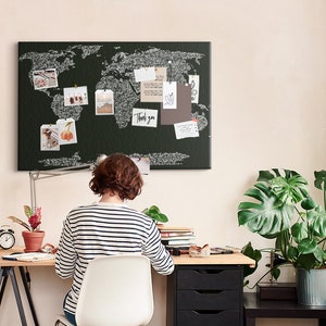 Peut inclure: Une personne est assise à un bureau avec un tableau en liège représentant une carte du monde en noir et blanc au-dessus d'elle. Le tableau en liège est décoré de photos, de notes et d'un mot "Merci". Le bureau est en bois et a un cadre en métal noir. Il y a une plante en pot sur le bureau et une plante en pot sur une table d'appoint.