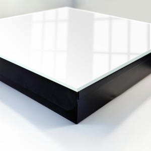 Può includere: Un espositore quadrato caratterizzato da una superficie superiore in vetro bianco liscio e riflettente, inserita in una solida base nera opaca. La struttura presenta bordi netti e definiti, con un design geometrico moderno e stabile.