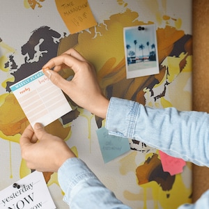 May include: A person is adding a weekly planner to a corkboard with a world map background. The planner has a blue and white design with the days of the week listed. There are other notes and a photo on the corkboard.