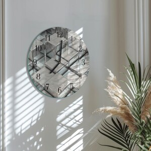 May include: A round wall clock with a glass face featuring a geometric design in shades of gray and white. The clock has black hands and numerals. The clock is mounted on a white wall, with shadows from a window visible.